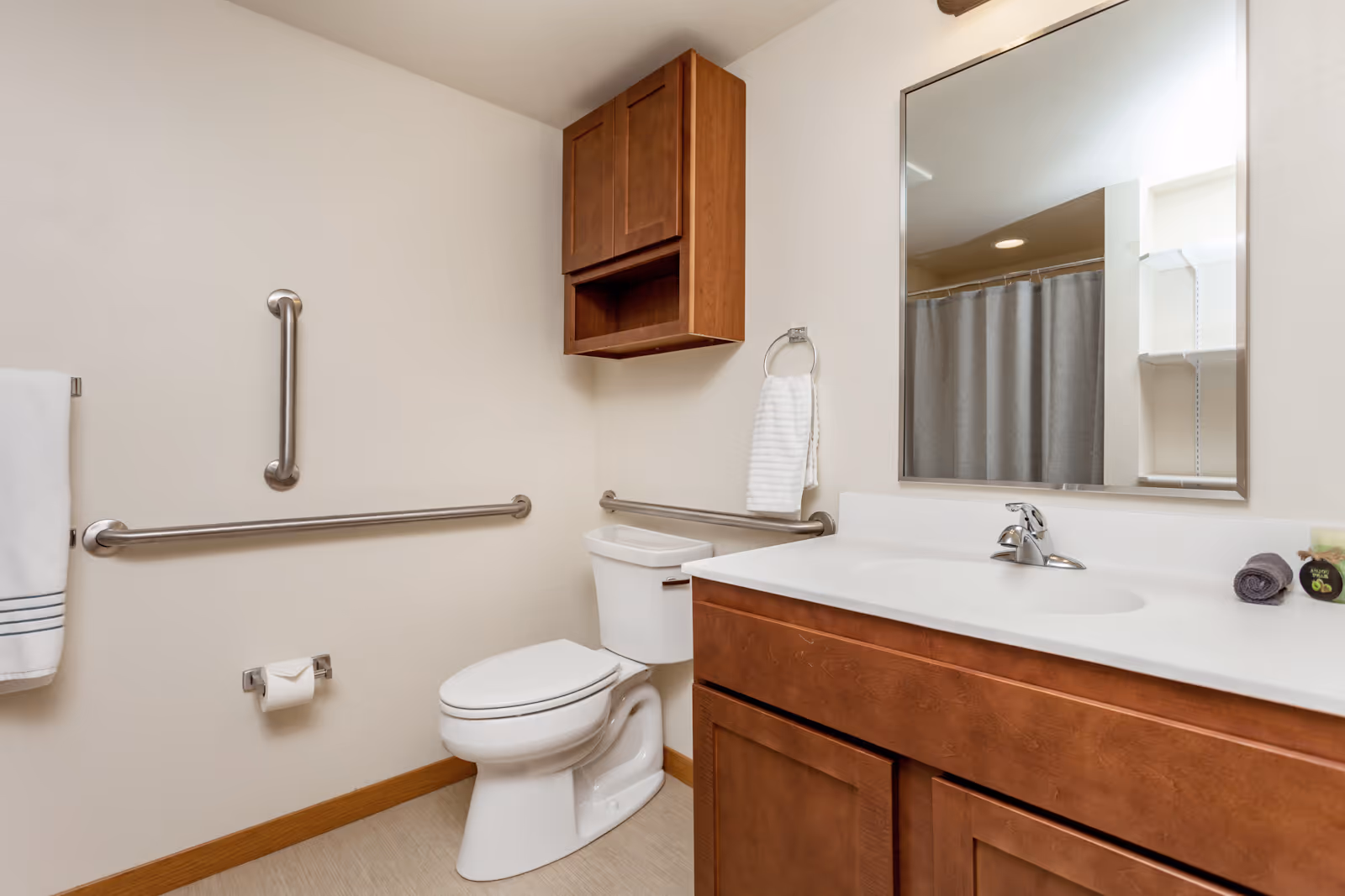 A clean and accessible bathroom featuring a white toilet with stainless steel grab bars on the walls, a wooden cabinet mounted above the toilet, a white countertop with a sink and faucet, a large mirror, and a towel ring holding a white hand towel. A shower curtain is partially visible in the mirror's reflection.