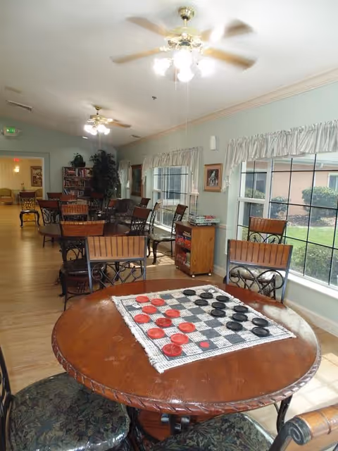 Bright common room with round tables and chairs, a checkerboard on the nearest table, large windows, ceiling fans, and a bookshelf in the background.