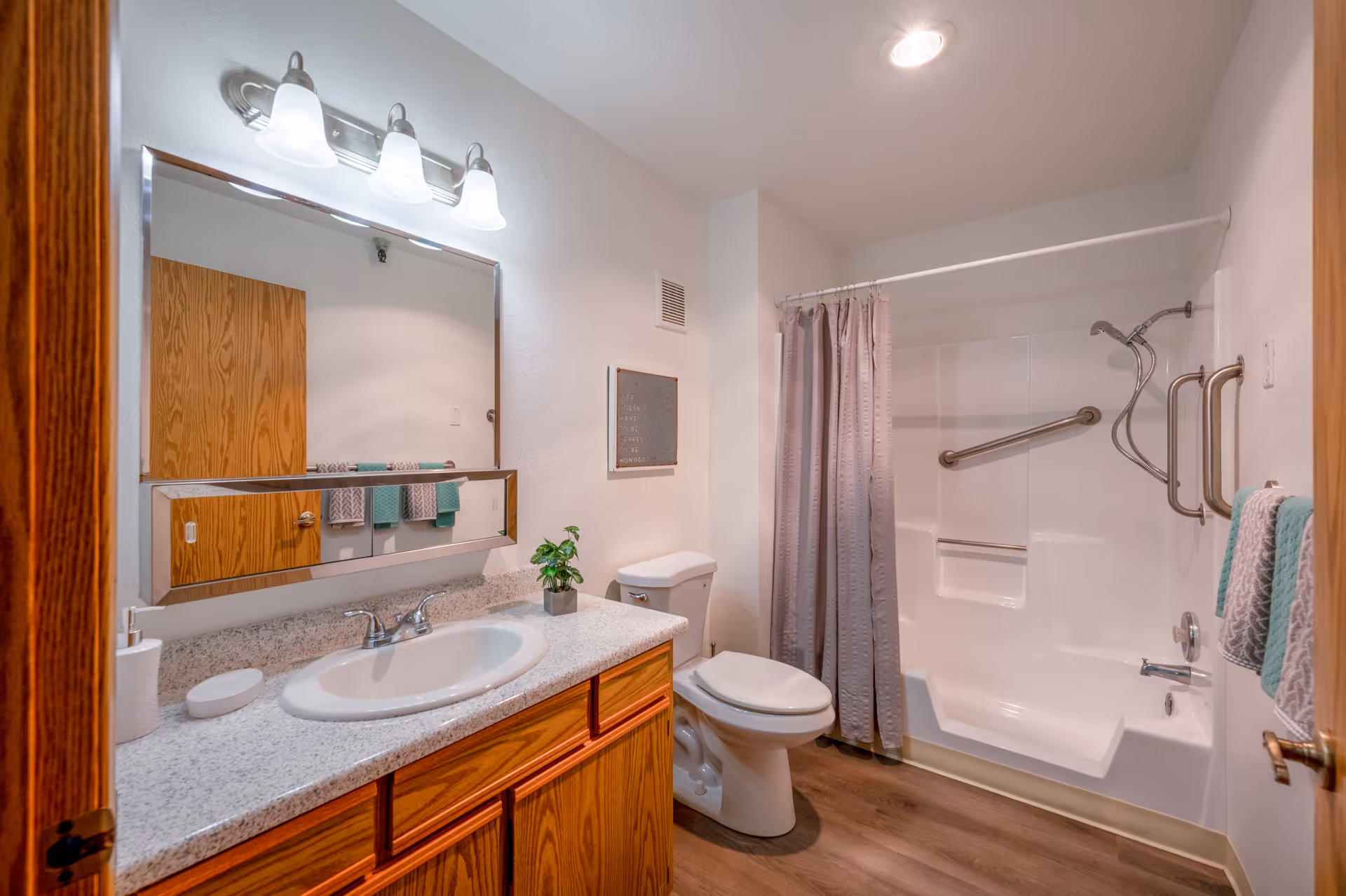 A clean bathroom featuring a white sink with a granite countertop, wooden cabinets, a large mirror with three overhead lights, a toilet, and a bathtub with a shower curtain and grab bars for accessibility. Towels hang on the wall next to the bathtub.
