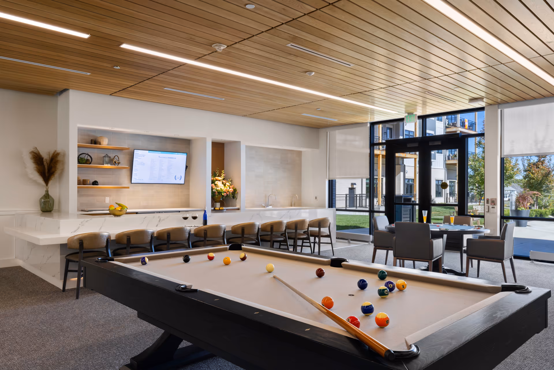A modern senior living facility common area featuring a pool table with billiard balls and cue sticks, a long marble countertop with bar stools, a wall-mounted TV, and a seating area with a round table and chairs near large windows that provide a view of the outdoor garden and buildings.