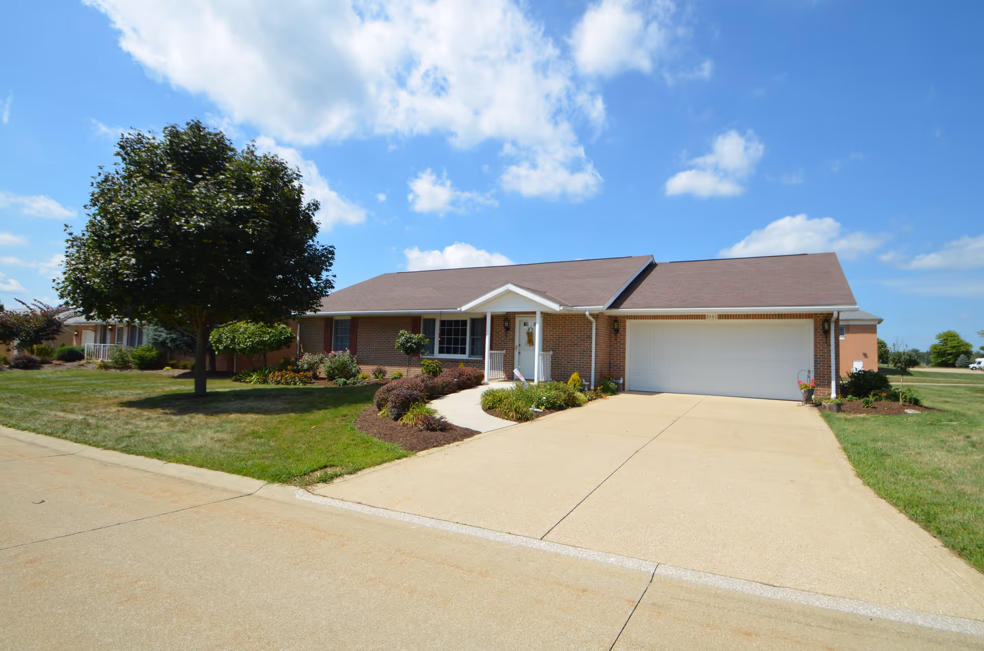 Single-story brick house with a two-car garage, a small front porch, and a well-maintained lawn with shrubs and a tree under a partly cloudy blue sky.
