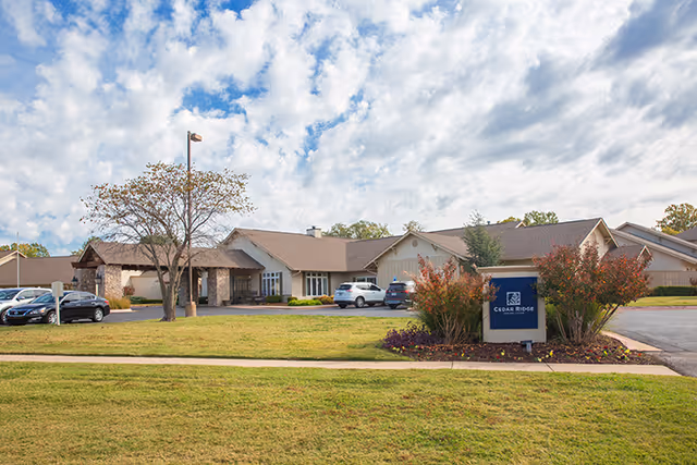 Exterior view of Cedar Ridge Senior Living facility showing a single-story building with a pitched roof, a parking area with several cars, a tree, and a sign with the facility's name surrounded by landscaping under a partly cloudy sky.