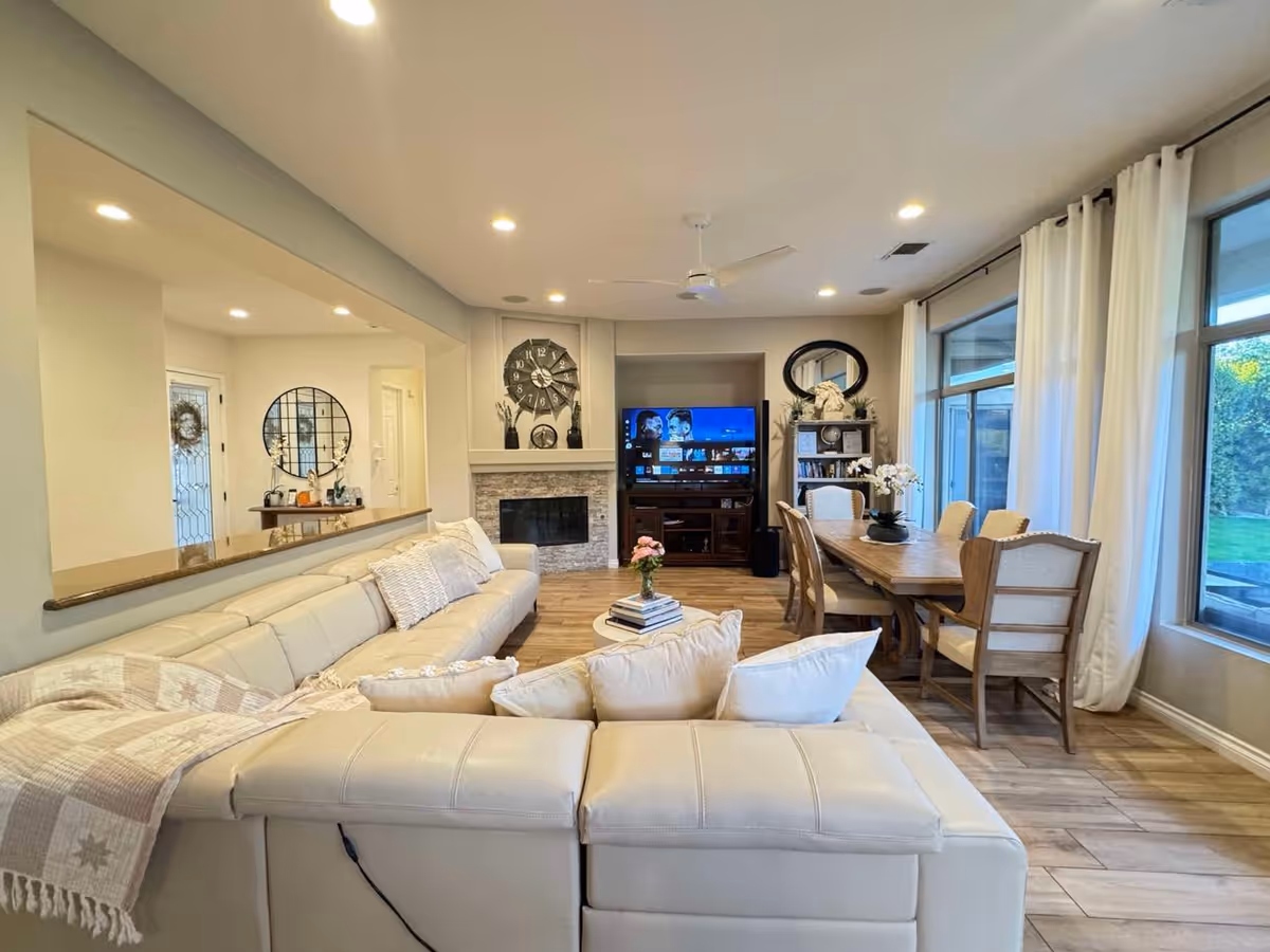 A bright and spacious living and dining area featuring a large cream-colored sectional sofa with pillows and a throw blanket, a wooden dining table with six upholstered chairs, a TV on a wooden stand, a fireplace with a decorative clock above it, large windows with white curtains letting in natural light, and wooden flooring.