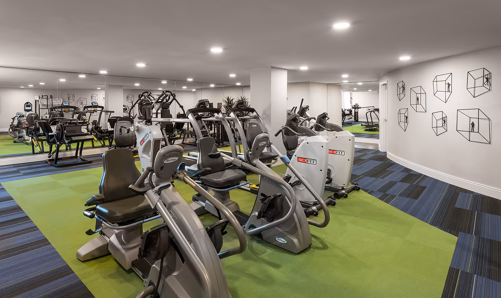 Bright indoor fitness center with cardio machines, mirrored wall, green and blue patterned flooring, and geometric wall art.