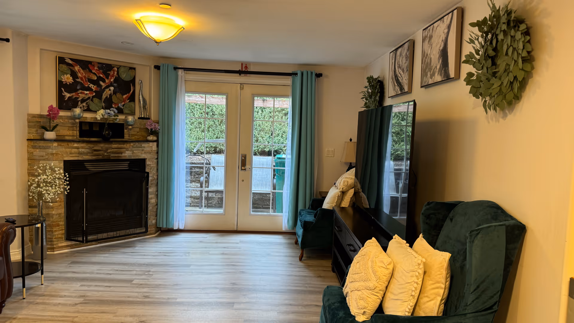 Living room with a stone fireplace on the left, French doors with turquoise curtains in the center, and green armchairs and a TV on the right.