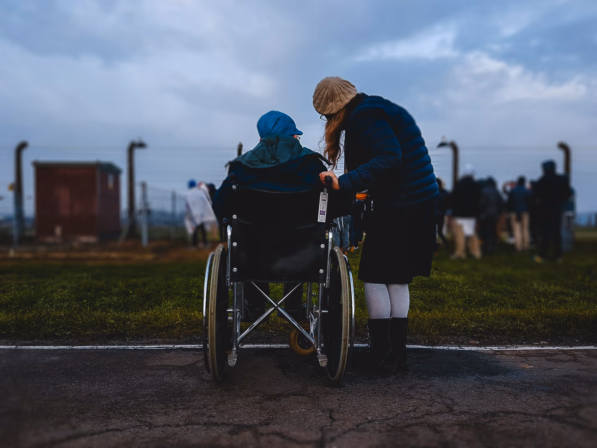 A person in a wheelchair wearing a blue jacket and hat is being pushed by another person dressed in a dark jacket, skirt, white tights, black boots, and a beige knit hat. They are outdoors on a paved path with grass on either side, and several other people are visible in the background near a fence under a cloudy sky.