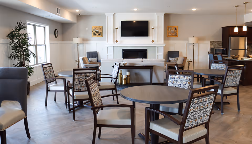 A bright and spacious senior living common area with several round tables and chairs arranged for socializing or dining. The room features a fireplace with a mounted flat-screen TV above it, two armchairs on either side, and a sofa facing the fireplace. There are floor lamps and decorative wall art, with a kitchen area visible in the background.