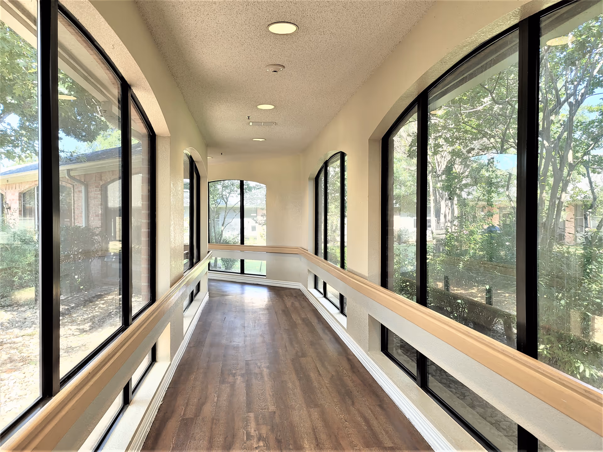 A bright hallway with large windows on both sides, allowing natural light to fill the space. The hallway has wooden flooring and beige walls with handrails along both sides. Outside the windows, trees and greenery are visible.