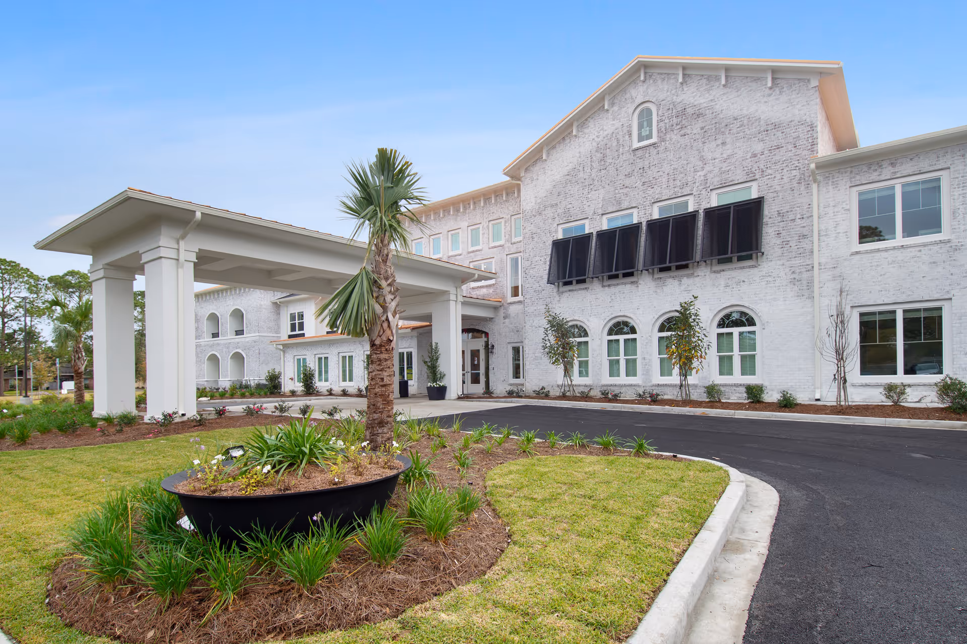 Front entrance of a white multi-story senior living building with a porte-cochère, circular driveway, and landscaped planter.