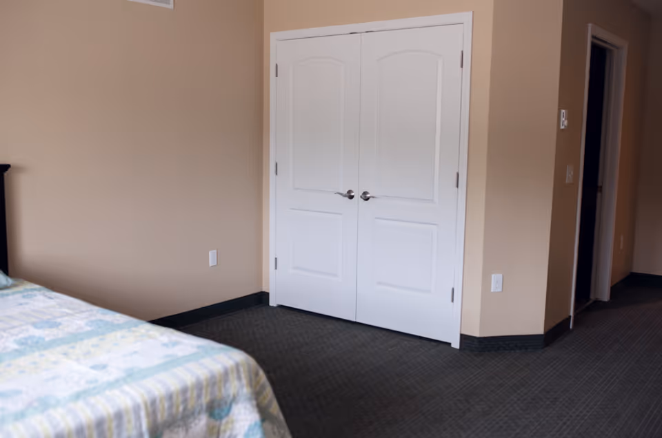 Corner of a bedroom showing a bed with a patterned quilt, beige walls, a double white closet door, and a hallway entrance.