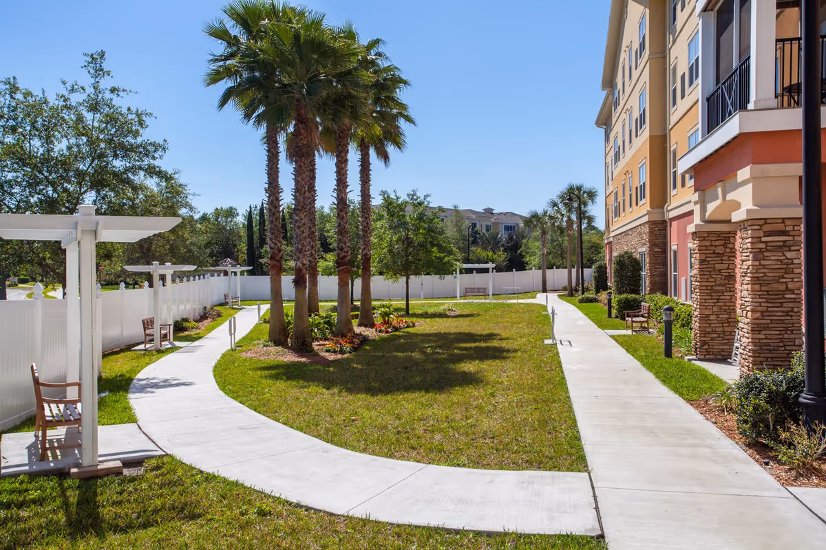 Outdoor garden area at Camellia At Deerwood with a curved concrete walkway, palm trees, benches, white fencing, and a multi-story building with stone pillars on the right side under a clear blue sky.