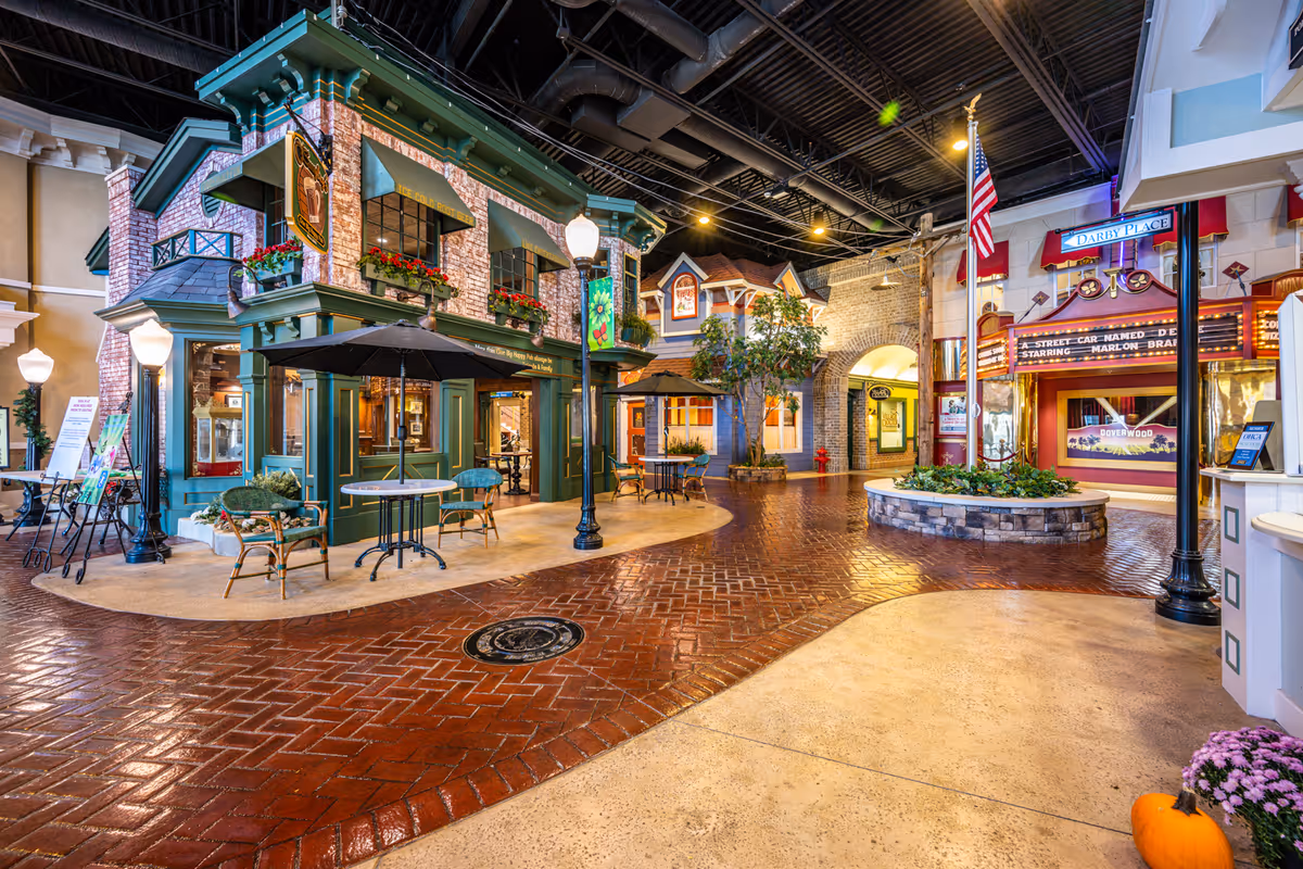 Indoor street-like setting in a senior living facility designed to resemble a small town with storefronts, outdoor-style seating, street lamps, and decorative plants under a high ceiling with exposed beams.