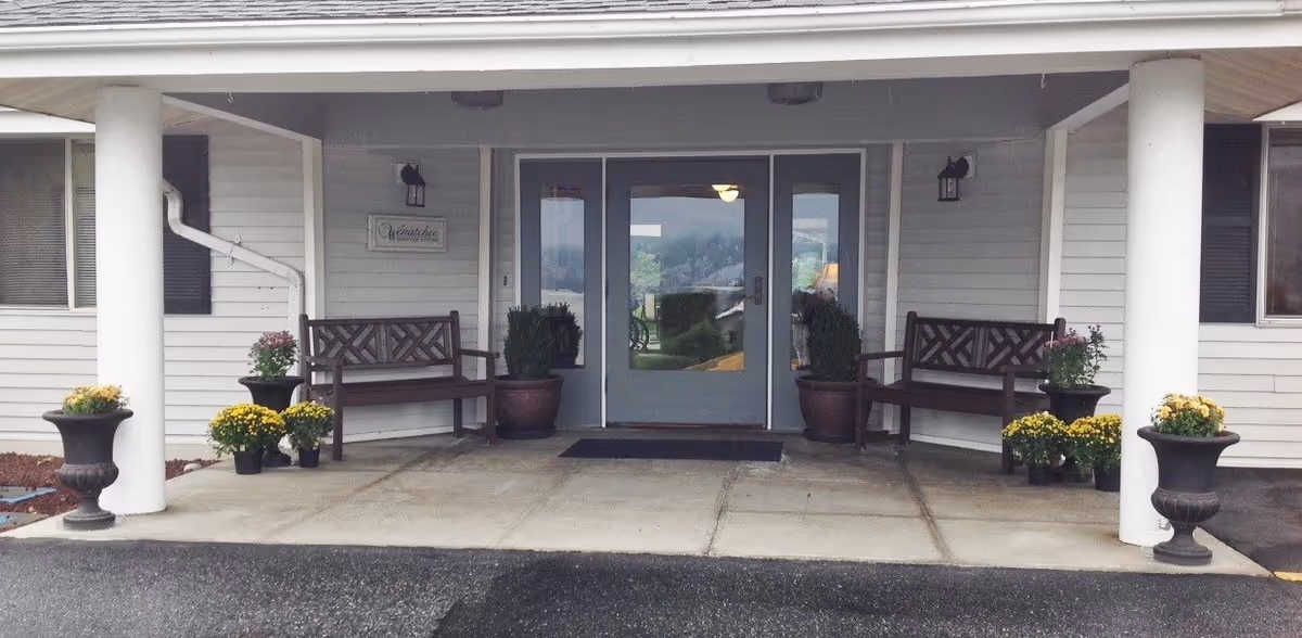 Entrance to a building with double glass doors, two wooden benches on either side, potted plants and flowers arranged around the benches, white pillars supporting a covered porch, and a sign on the wall that reads 'Wreathe'.