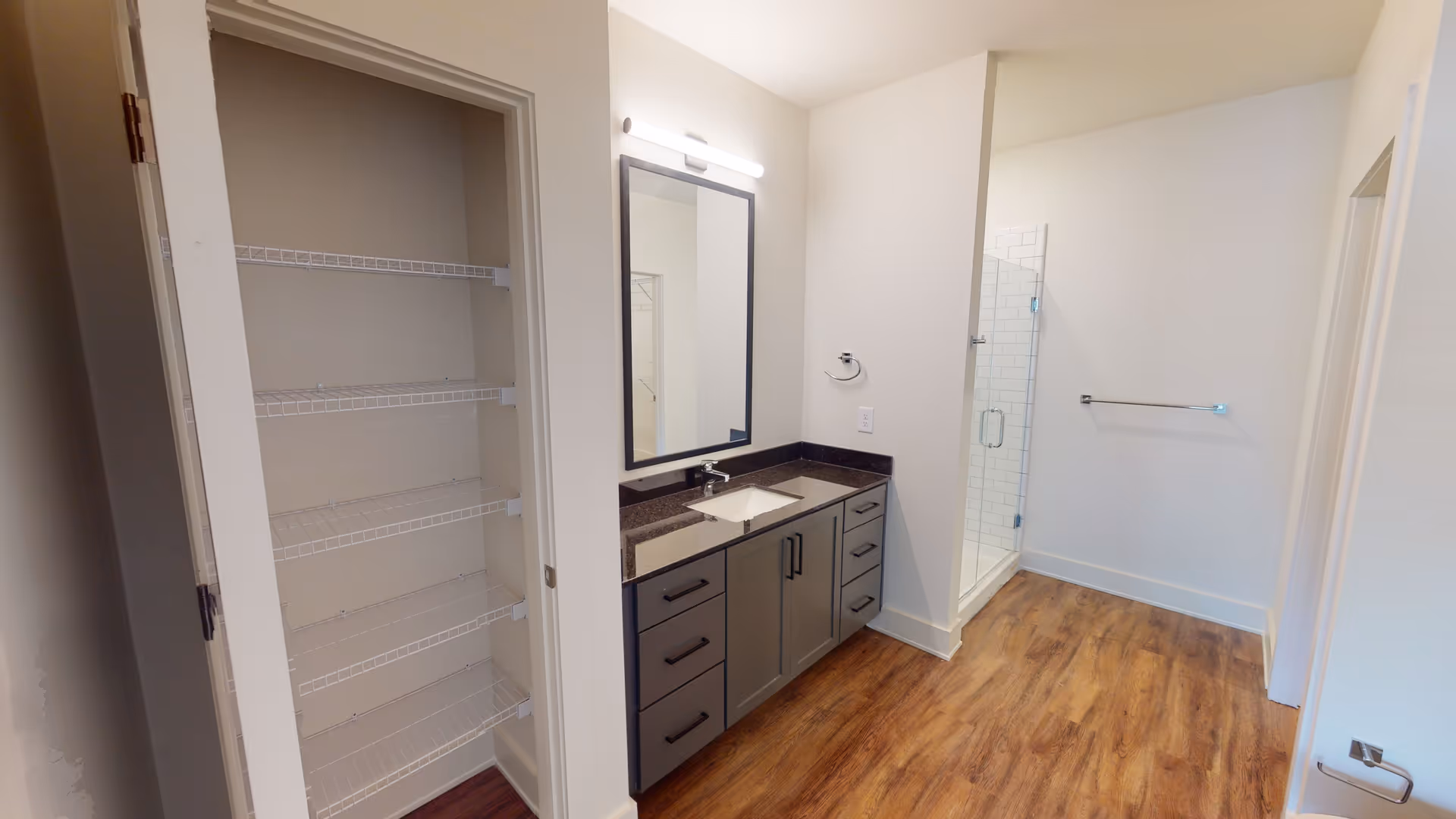 A modern bathroom featuring a dark countertop vanity with multiple drawers and cabinets, a rectangular mirror with a light above it, a glass-enclosed shower with white subway tiles, a towel rack on the wall, and a closet with wire shelving on the left side. The floor is wood-style laminate.