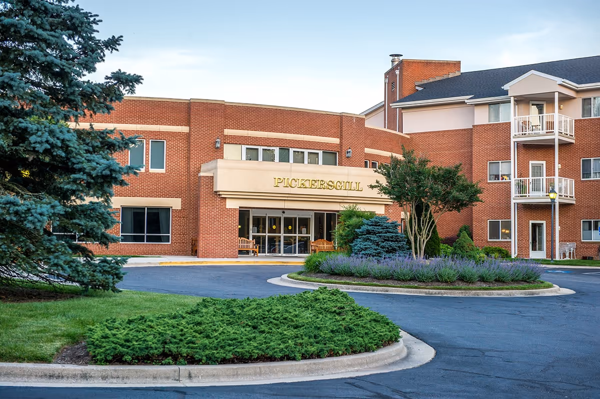 Exterior view of Pickersgill Retirement Community building with a circular driveway, landscaped greenery including bushes and trees, and a clear sky.