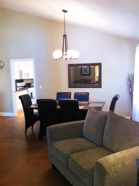 Interior view of a living and dining area with a beige sofa in the foreground, a dining table with six black chairs, a hanging light fixture above the table, a large mirror on the wall, and a doorway leading to a kitchen with white cabinets and appliances.