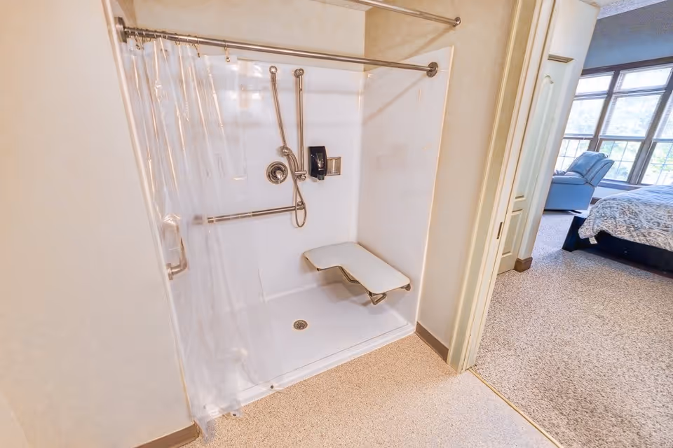 A walk-in shower with a clear plastic curtain, a fold-down shower seat, and grab bars. The shower has a handheld showerhead and a soap dispenser. The shower area opens into a carpeted bedroom with a bed and recliner chair near large windows.