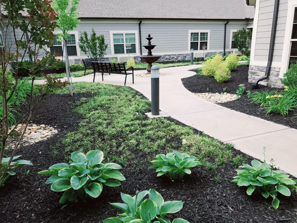 Outdoor garden area at Morning Pointe of Owensboro featuring a concrete walkway, green plants, shrubs, a black metal bench, and a multi-tiered water fountain in front of a light gray building with white-trimmed windows.