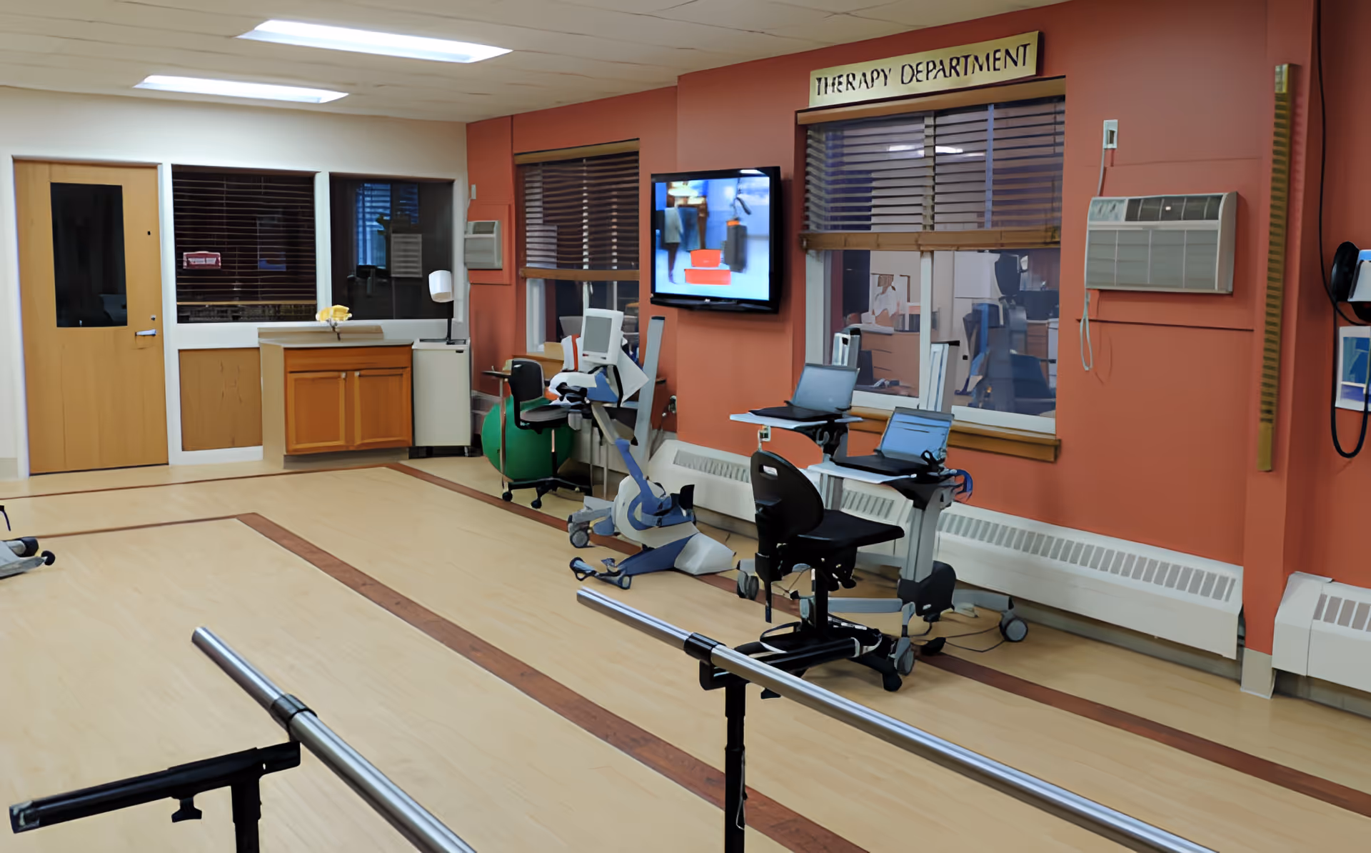 Rehabilitation therapy room with parallel bars, exercise equipment, chairs and monitors against a coral-colored wall.