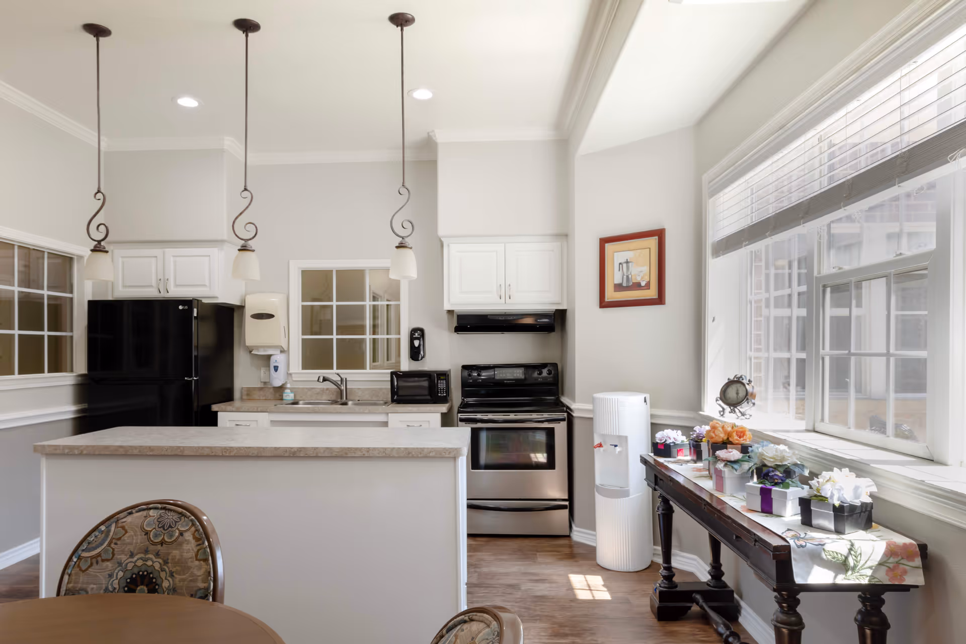 A bright kitchen area with white cabinets, a black refrigerator, a black stove, and a microwave. There are three pendant lights hanging from the ceiling above a countertop. A table with floral-patterned chairs is partially visible in the foreground. A side table near a large window holds several small potted flowers and a decorative clock. The room has light-colored walls and wood flooring.
