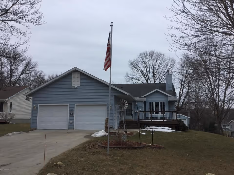A single-story blue house with a double garage, a front porch with railing, and an American flag on a flagpole in the front yard. The yard has patches of snow and leafless trees surrounding the property under a cloudy sky.