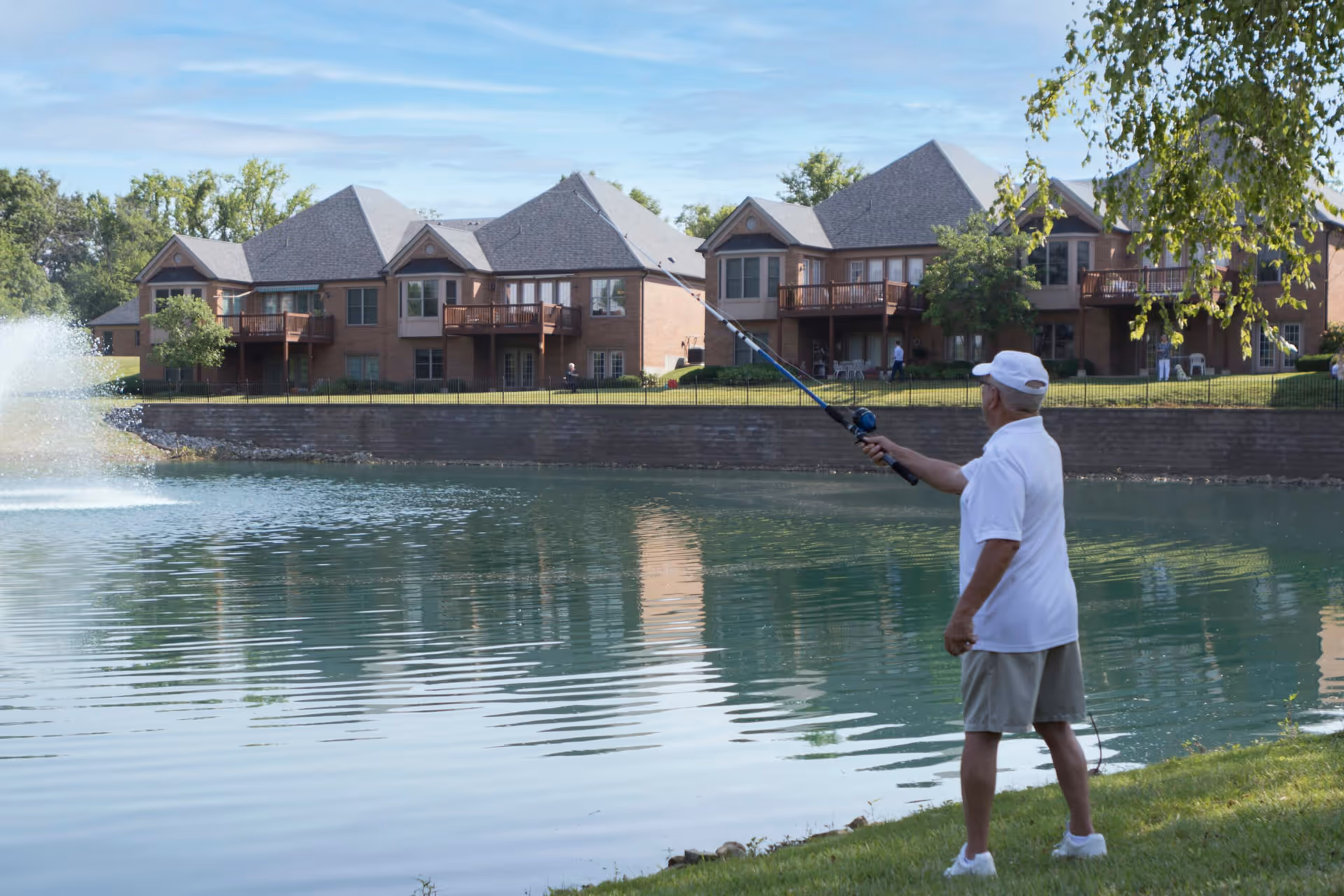 An elderly man wearing a white cap, white shirt, and beige shorts is fishing by a pond with a fountain. In the background, there are large brick residential buildings with balconies and green lawns under a partly cloudy sky.