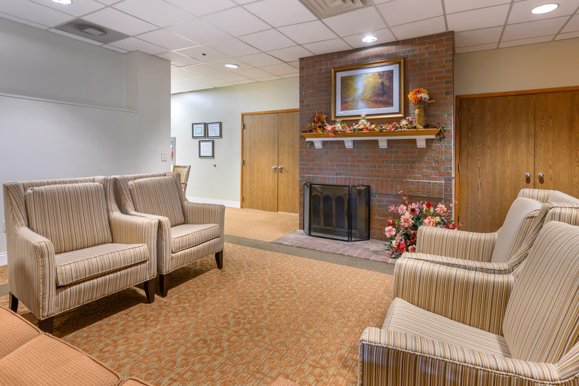 A cozy sitting area with four striped armchairs arranged around a brick fireplace. The fireplace mantel is decorated with autumn-themed garlands and a vase with flowers, and above it hangs a framed painting of a forest scene. The room has a carpeted floor and wooden doors in the background.