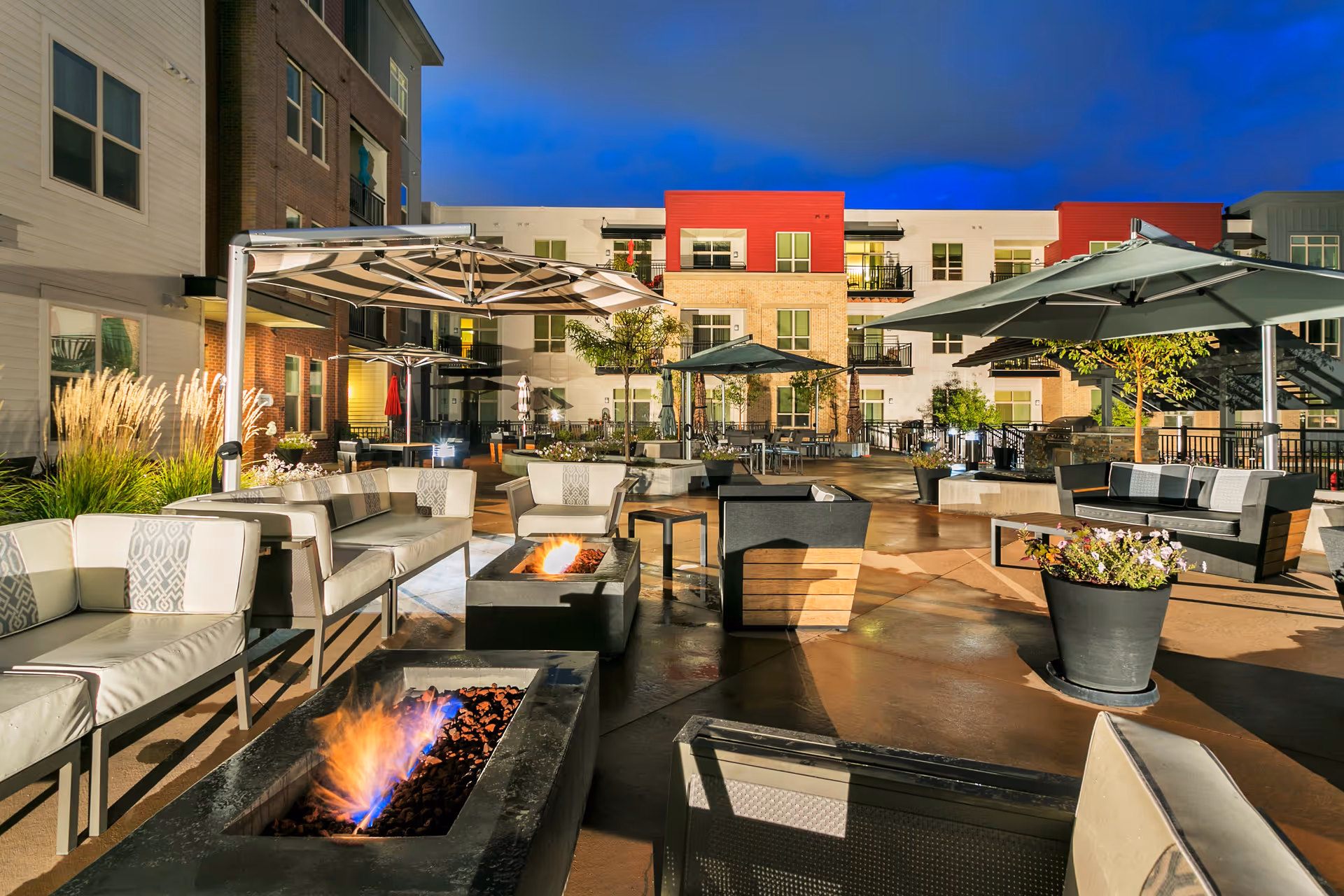 Outdoor patio area at dusk with modern seating arrangements, fire pits, large umbrellas, potted plants, and a multi-story residential building in the background.
