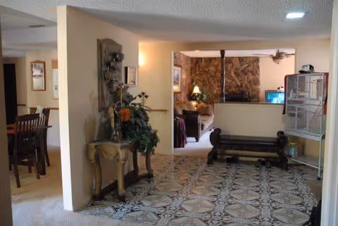 Interior view of a senior living facility showing a hallway with patterned tile flooring leading to a living room area with a stone wall and a ceiling fan. To the left, there is a dining area with a wooden dining table and chairs. A decorative table with flowers and a wall-mounted item is on the left side of the hallway. A bench and a large birdcage are on the right side of the hallway.