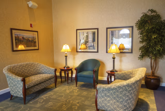 A cozy assisted-living sitting area with upholstered chairs and loveseats, side tables with lamps, framed artwork, and a potted plant.