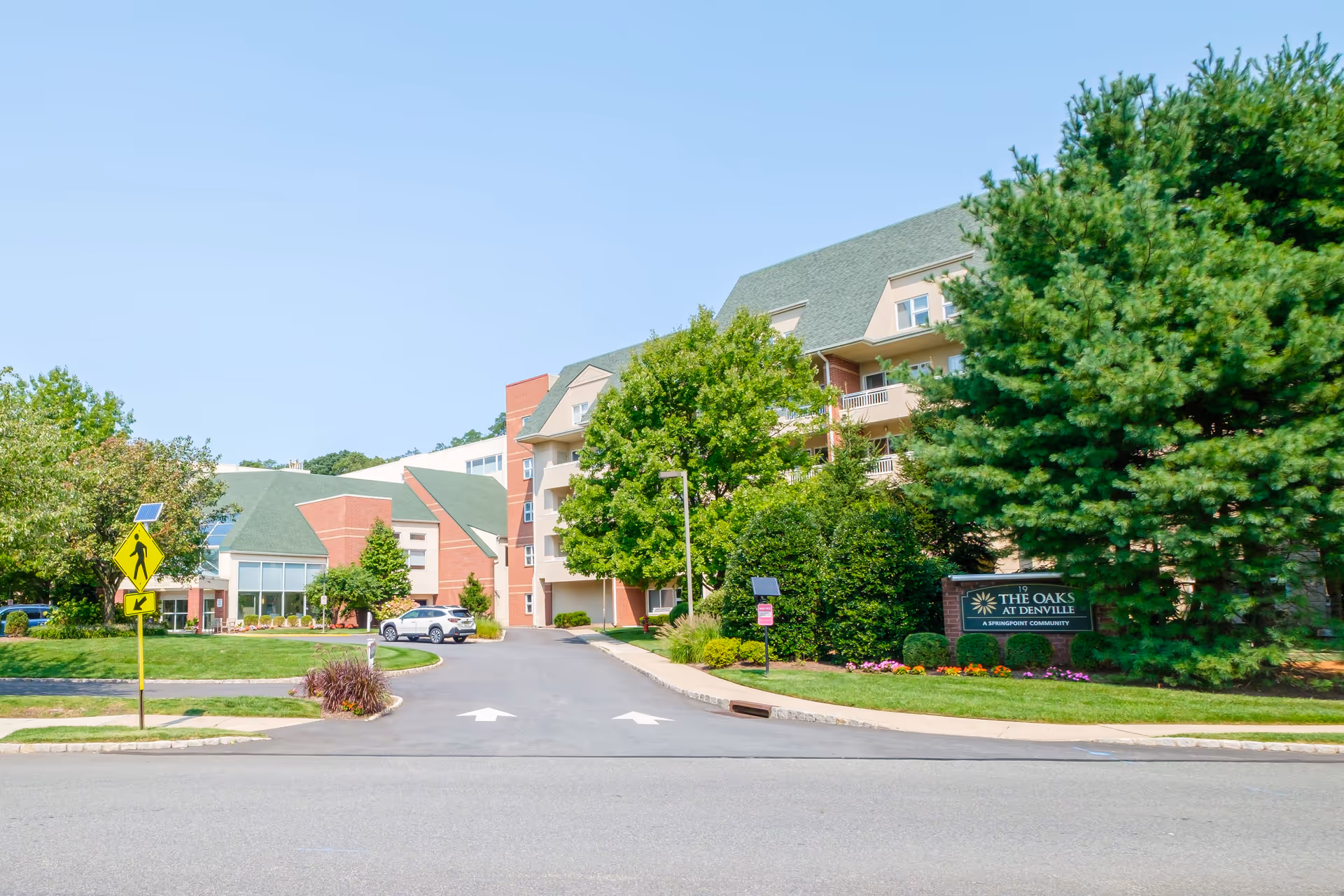 Exterior view of The Oaks At Denville senior living facility showing a multi-story building with green roofs, surrounded by trees and landscaping. A driveway leads into the facility with a pedestrian crossing sign visible on the left. The facility sign is visible on the right side near the entrance.