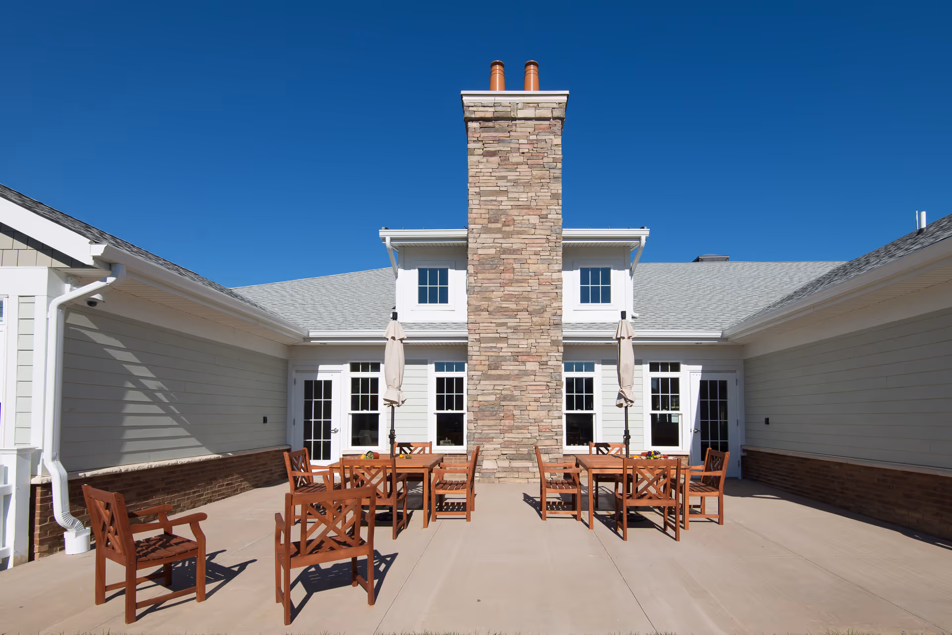 Outdoor patio area at The Terraces at Lawrence featuring wooden tables and chairs with closed umbrellas, centered around a tall stone chimney attached to a light-colored building under a clear blue sky.