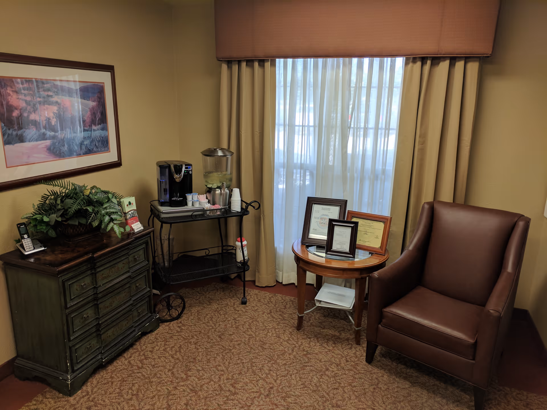 A cozy interior sitting area with a leather armchair, round side table holding framed certificates, a beverage cart, dresser with plants, and a curtained window.
