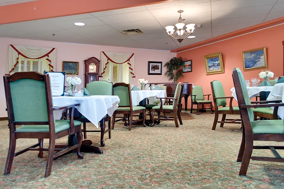 A dining room in a senior living facility with several tables covered in white tablecloths and set with glassware and floral centerpieces. The room has green cushioned wooden chairs, a patterned carpet, peach-colored walls adorned with framed paintings, and windows with decorative curtains. A chandelier hangs from the ceiling, and a grandfather clock is visible against the far wall.