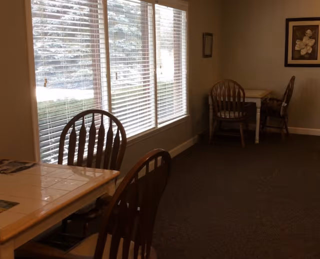 A quiet interior room with several wooden chairs and tables arranged near large windows with white blinds. The room has beige walls, carpeted floor, and a framed flower picture on the wall.