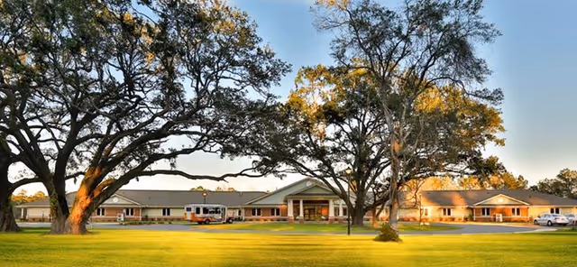 Single-story assisted living facility building with large oak trees and a well-kept lawn in front.