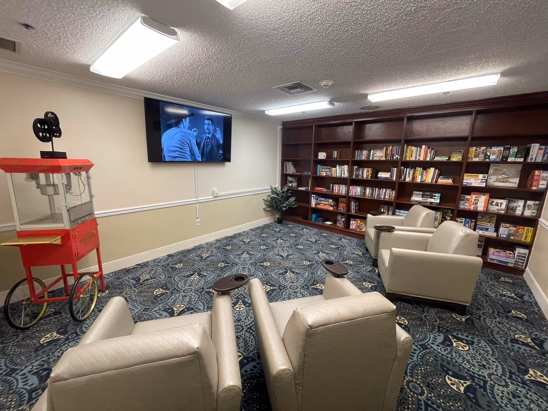 Small media lounge with four beige recliners facing a wall-mounted TV, bookshelves along the back wall, and a red popcorn cart.