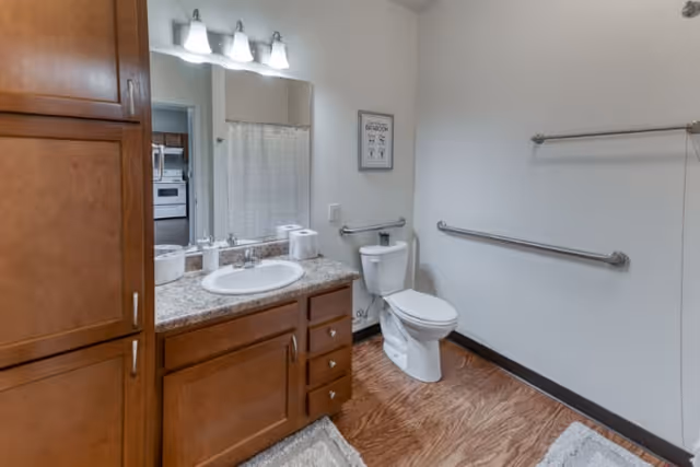 A clean bathroom featuring a wooden vanity with a granite countertop, a white sink, and a large mirror above it. There are three light fixtures above the mirror. The bathroom has a white toilet with grab bars on the adjacent walls for accessibility. A towel rack is mounted on the right wall, and a framed sign hangs above the toilet. The floor has a wood-like finish, and a small rug is placed in front of the vanity. A doorway leads to a kitchen area visible in the background.