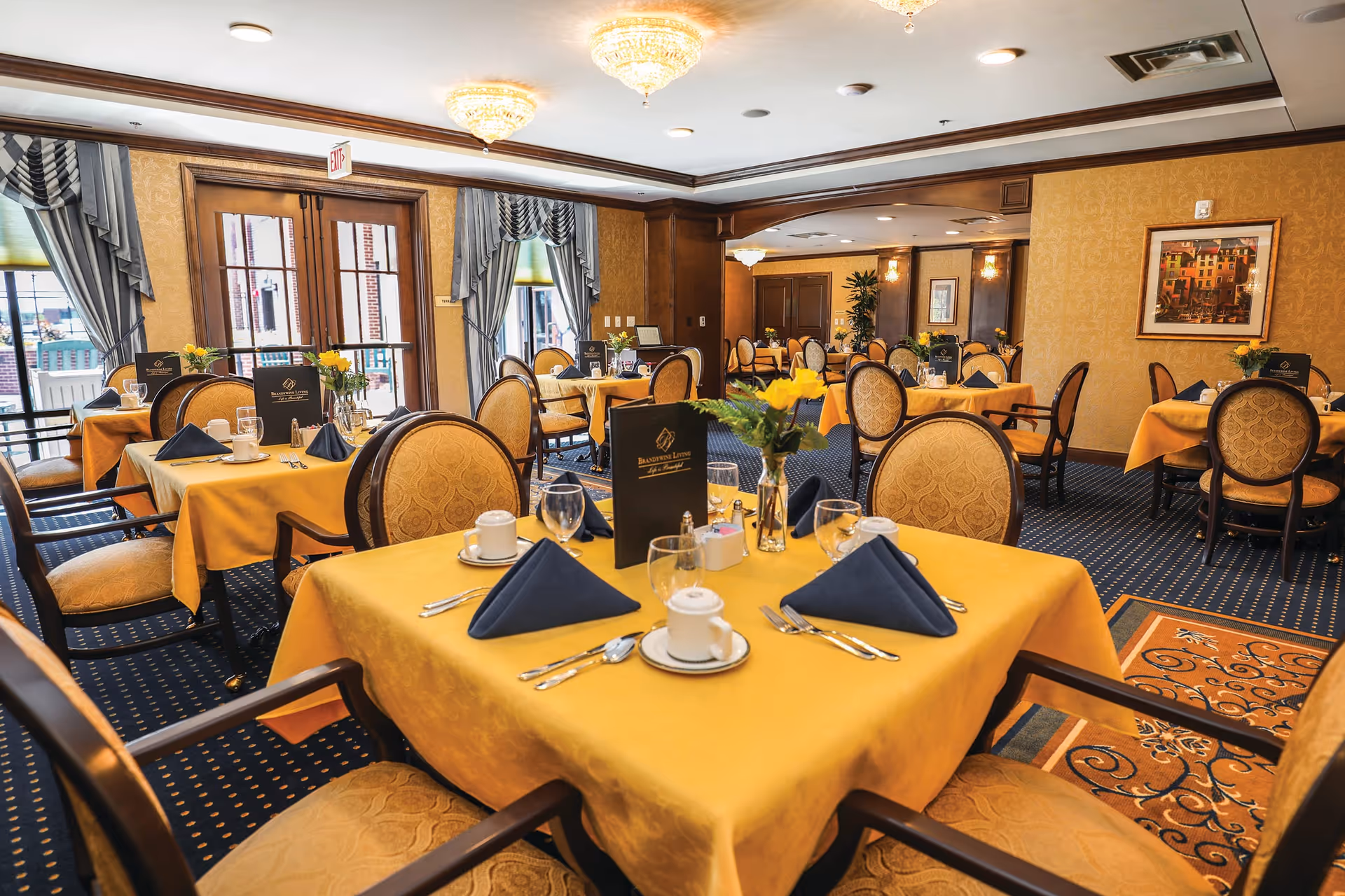 A dining room with multiple tables covered in yellow tablecloths, each set with blue folded napkins, cups, glasses, and silverware. The room features upholstered chairs with patterned fabric, large windows with draped curtains, and warm lighting from ceiling fixtures. There are flower arrangements on each table and framed artwork on the walls.