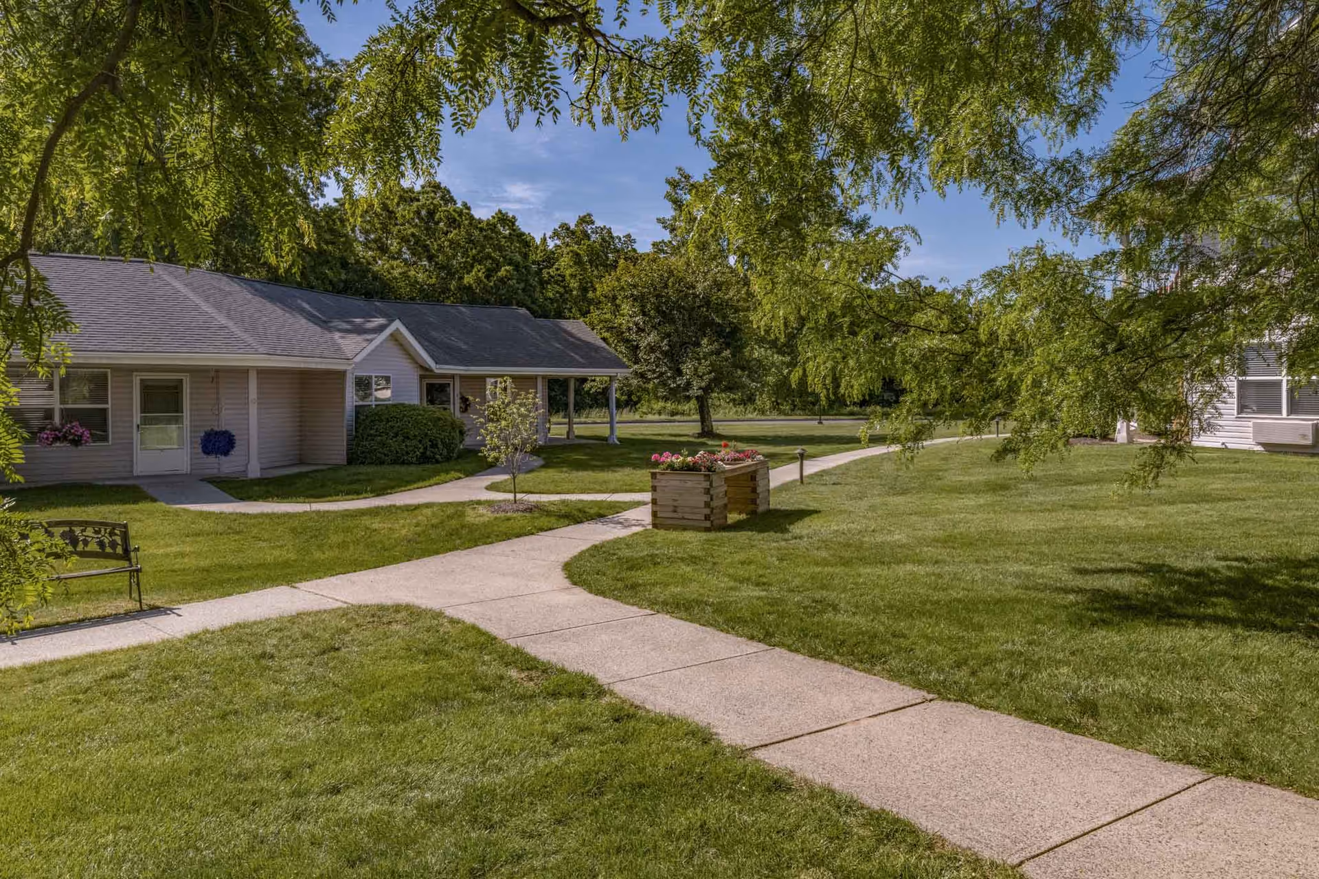 A peaceful outdoor area at Holiday Quail Run Estates featuring a paved walkway winding through well-maintained green lawns, a wooden planter box with colorful flowers, a bench, and single-story residential buildings surrounded by trees under a clear blue sky.
