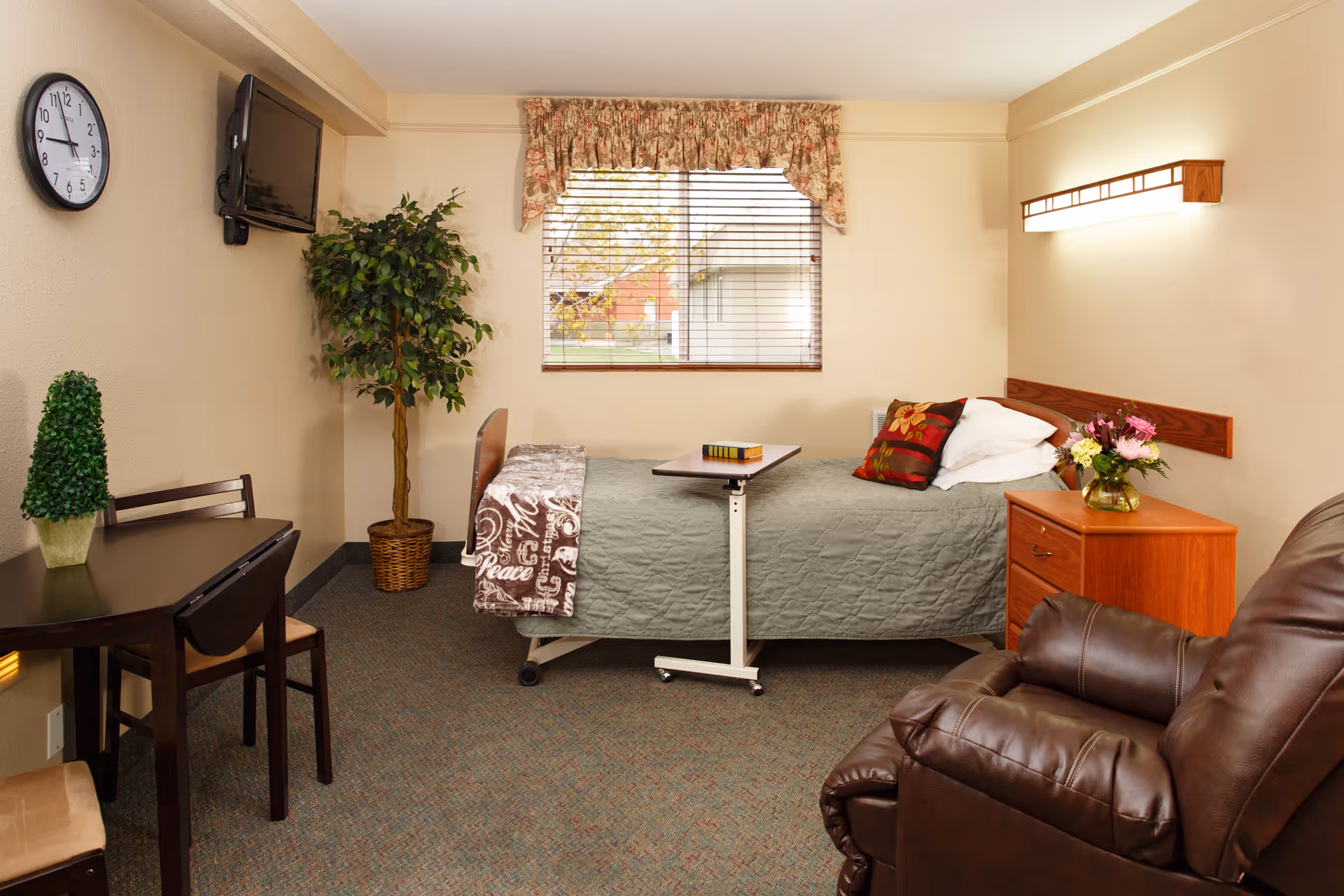 A bright senior living bedroom with a single bed, overbed table, nightstand with flowers, recliner, small dining table and wall-mounted TV.