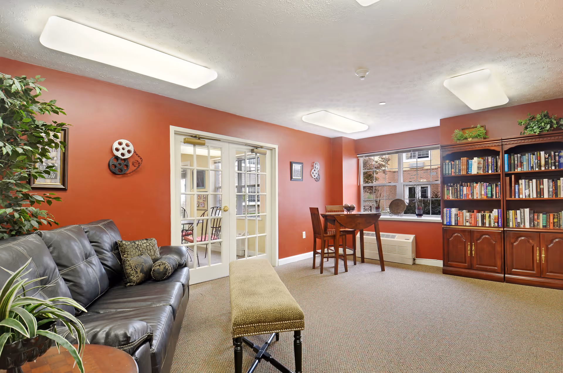 A cozy senior living common area with red walls, a black leather couch with decorative pillows, a green cushioned bench, a wooden table with two chairs near a window, and a large wooden bookshelf filled with books. There are plants and wall decorations, and a set of glass double doors leading to another room.