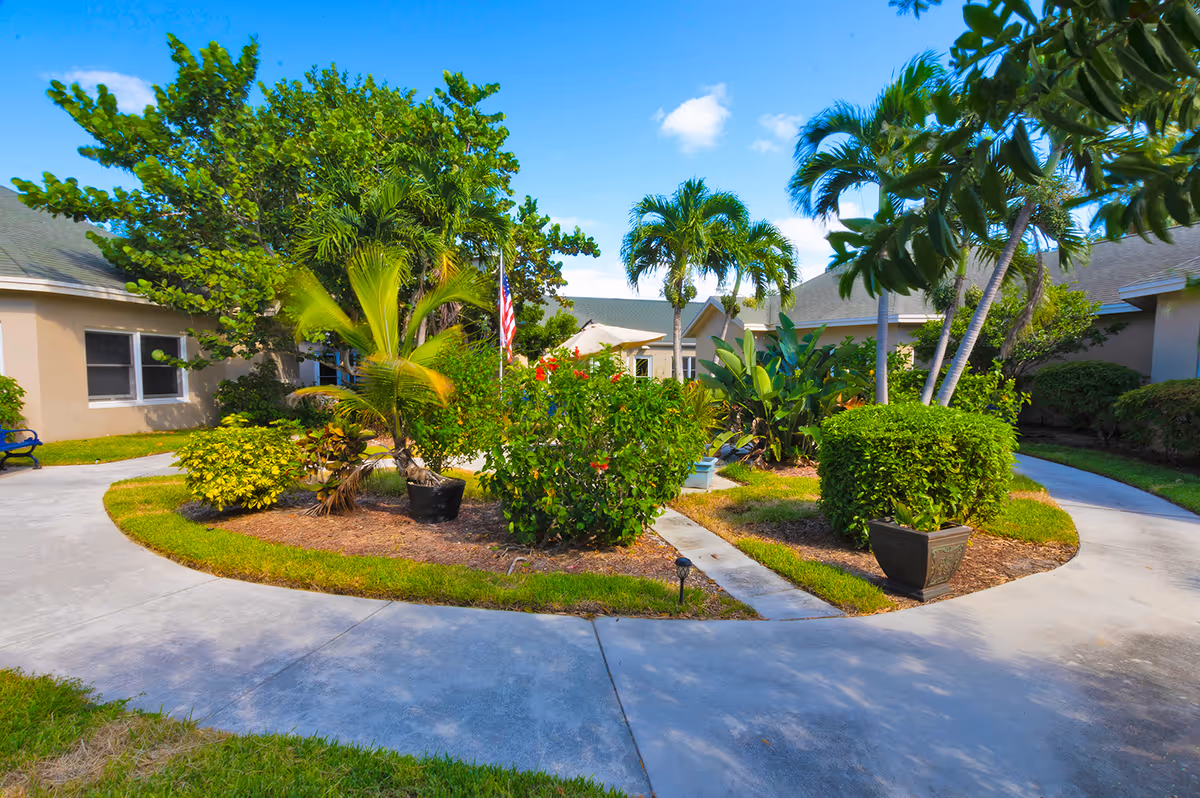 A landscaped courtyard area with a curved concrete pathway surrounding a garden bed filled with various tropical plants and shrubs, including palm trees. Beige buildings with windows and a green roof surround the courtyard under a blue sky with a few clouds.