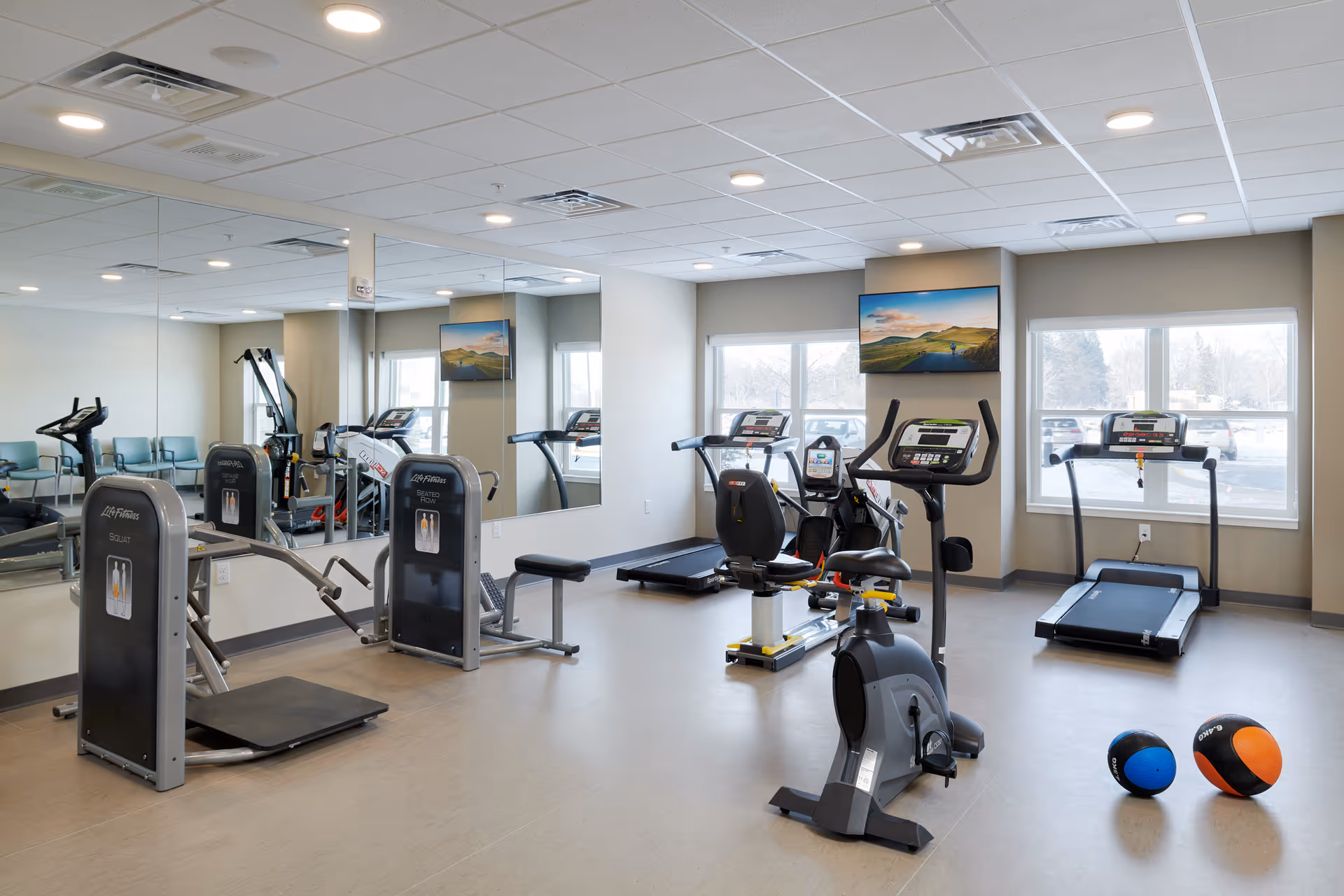Bright fitness room with treadmills, stationary bikes, strength machines, wall mirrors, and medicine balls.