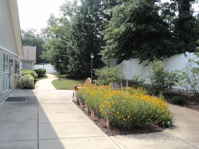 Outdoor walkway beside a building with a garden bed filled with yellow flowers and small American flags. There are trees and bushes along a white fence in the background, and a bench is visible near the garden bed.