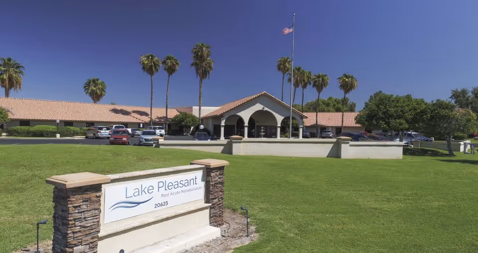 Exterior view of Lake Pleasant Post Acute Rehabilitation Center with a large green lawn in front, palm trees, a flagpole with an American flag, and a parking lot with several cars. The building has a tiled roof and an arched entrance.