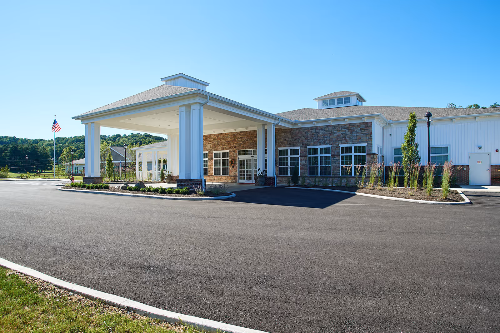 Front entrance of a single-story senior living building with a covered porte-cochere, landscaped beds, and an American flag under a clear blue sky.
