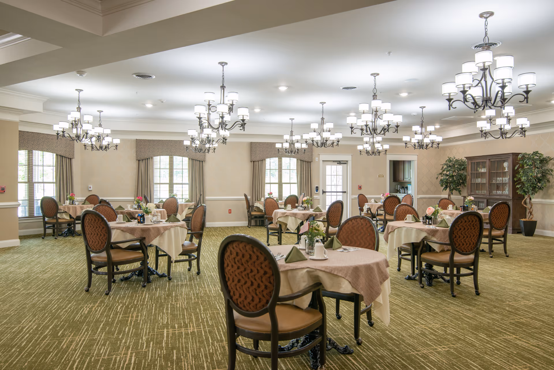 Large dining room with round tables covered in tablecloths, upholstered chairs, chandeliers, and place settings.