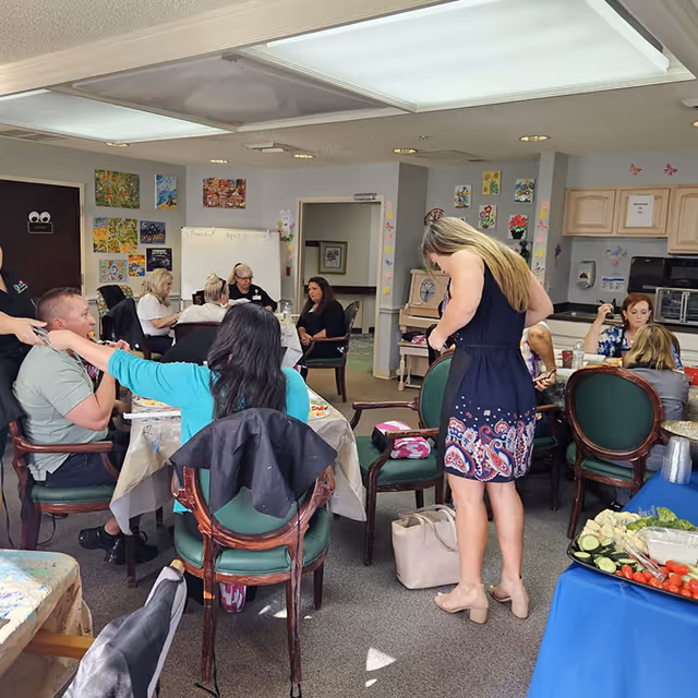 Several people gather around tables in a communal activity room with artwork on the walls and a buffet table of vegetables.