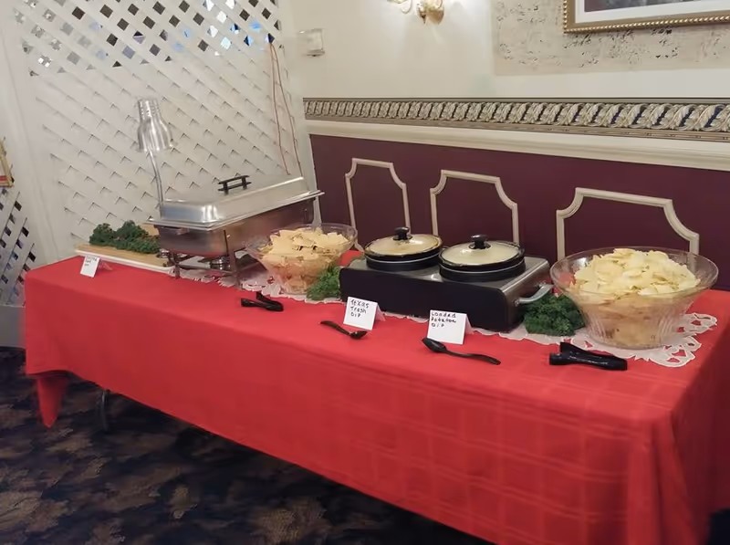 A buffet table covered with a red tablecloth displaying a food warmer, two bowls of chips, two covered pots labeled Texas Trash Dip and Loaded Potato Dip, and some greenery for decoration. The background includes a white lattice and a wall with decorative molding and a framed picture.